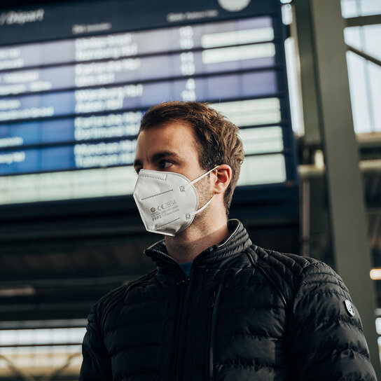 Mann mit FFP2 Maske vor Anzeigetafel in Bahnhof
